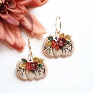 Pode incluir: Um par de brincos bege em forma de ab&oacute;bora com argolas douradas. Cada brinco &eacute; adornado com um arranjo floral de flores vermelhas, brancas e amarelas e folhas verdes. Uma grande flor rosa ao fundo.