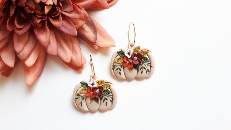 Pode incluir: Um par de brincos bege em forma de ab&oacute;bora com argolas douradas. Cada brinco &eacute; adornado com um arranjo floral de flores vermelhas, brancas e amarelas e folhas verdes. Uma grande flor rosa ao fundo.