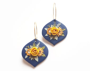 May include: A pair of blue teardrop-shaped earrings with gold hoops. Each earring features a yellow rose and other floral details in shades of yellow, orange, and green.