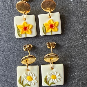 May include: Two pairs of drop earrings featuring gold-tone circular studs attached to square white pendants. The top pair displays yellow daffodils with orange centers, while the bottom pair features white daisies with yellow centers and green stems.