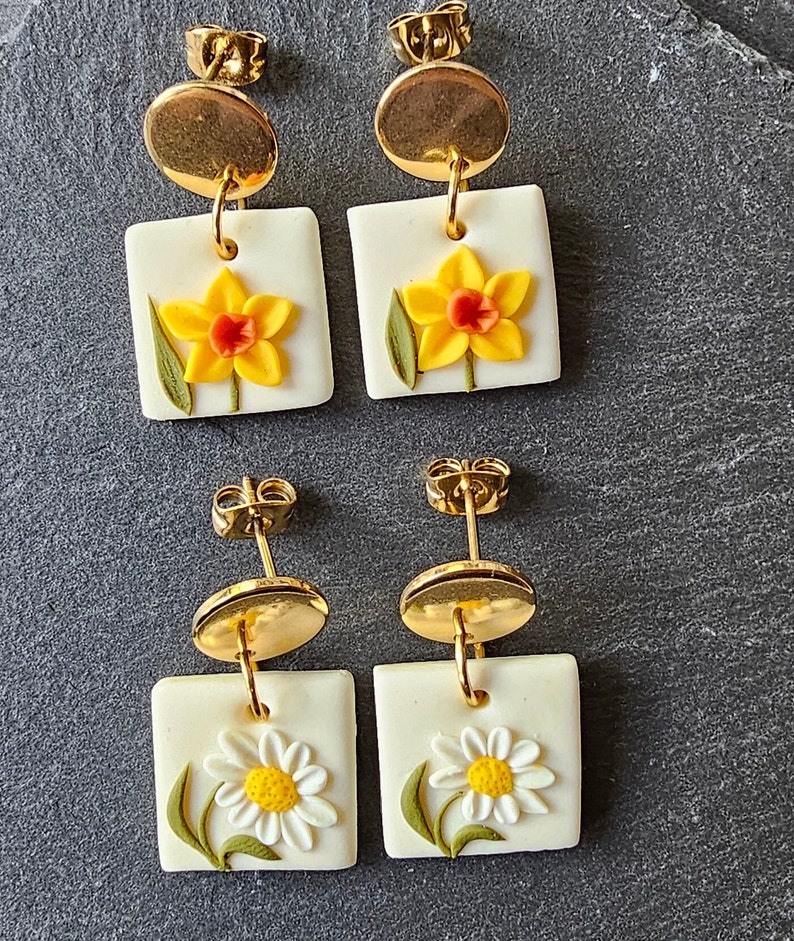 May include: Two pairs of drop earrings featuring gold-tone circular studs attached to square white pendants. The top pair displays yellow daffodils with orange centers, while the bottom pair features white daisies with yellow centers and green stems.
