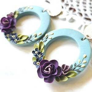 Könnte beinhalten: Ein Paar hellblaue, kreisförmige Ohrringe mit dreidimensionalen violetten Rosen, kleinen dunkelblauen Blüten und hellgrünen Blättern am unteren Teil jedes Reifens. Die Ohrringe haben eine glatte, matte Oberfläche und metallische Befestigungen.