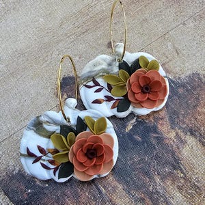 May include: A pair of white pumpkin-shaped earrings with gold-colored hoops. Each earring features a 3D floral design with orange, green, and black accents. The pumpkins have a marbled effect and are decorated with leaves.
