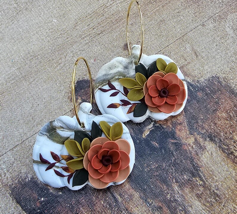 May include: A pair of white pumpkin-shaped earrings with gold-colored hoops. Each earring features a 3D floral design with orange, green, and black accents. The pumpkins have a marbled effect and are decorated with leaves.