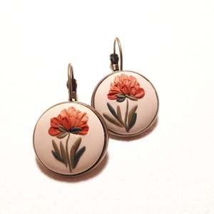May include: A pair of round drop earrings featuring a light beige background with a raised, sculpted orange flower and dark green leaves. The circular design is set in a bronze-toned metal frame with a matching lever-back closure mechanism.