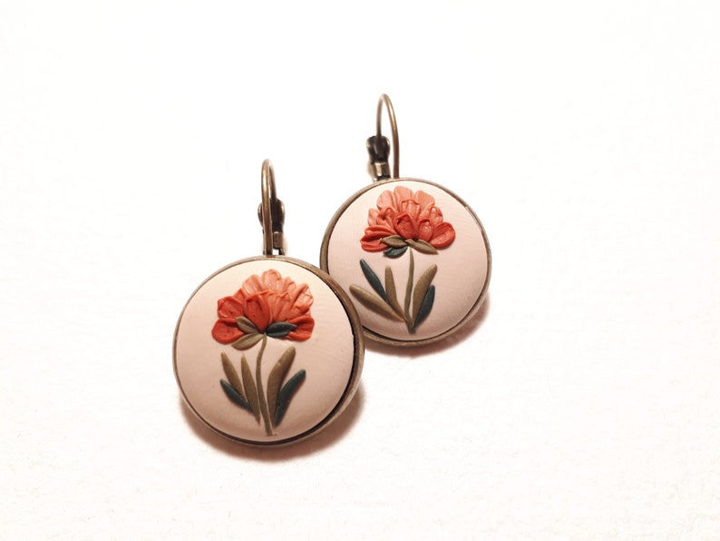 May include: A pair of round drop earrings featuring a light beige background with a raised, sculpted orange flower and dark green leaves. The circular design is set in a bronze-toned metal frame with a matching lever-back closure mechanism.