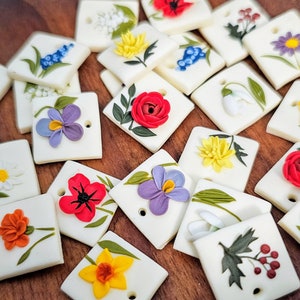 May include: A collection of square, off-white ceramic tiles, each featuring a raised, three-dimensional floral design in various colors including red, yellow, purple, orange, and blue, with a small circular hole punched near one corner.