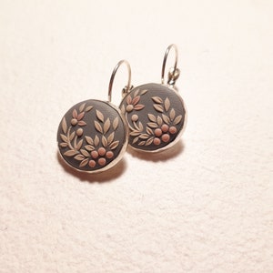Peut inclure: Une paire de boucles d'oreilles rondes avec une base gris fonc&eacute; orn&eacute;e d'un relief de feuilles beige clair et de petites baies roses. Les disques circulaires sont sertis dans un cadre en m&eacute;tal argent&eacute; et suspendus &agrave; des attaches dormeuses.