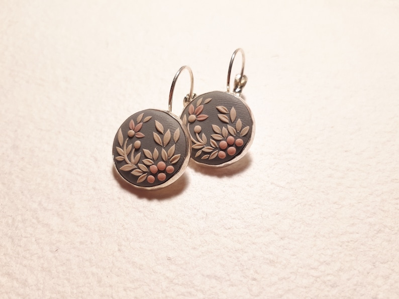 Peut inclure: Une paire de boucles d'oreilles rondes avec une base gris fonc&eacute; orn&eacute;e d'un relief de feuilles beige clair et de petites baies roses. Les disques circulaires sont sertis dans un cadre en m&eacute;tal argent&eacute; et suspendus &agrave; des attaches dormeuses.