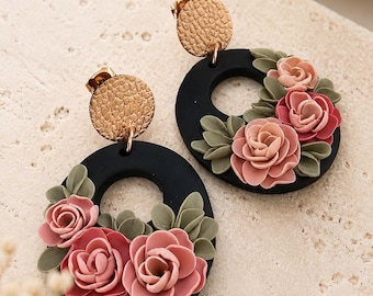 May include: A pair of dangle earrings featuring a circular black base adorned with sculpted pink roses and sage green leaves. Each earring is attached to a textured gold-toned circular stud with a post back closure, creating a floral design.