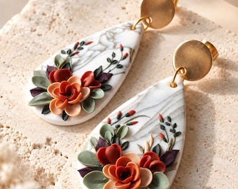 May include: A pair of teardrop-shaped earrings featuring a white marble-like pattern with raised, three-dimensional floral embellishments in shades of burnt orange, peach, and sage green, attached to circular, brushed gold-tone metal studs.