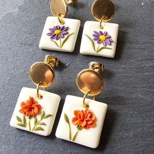 May include: Two pairs of drop earrings featuring gold-tone circular studs attached to white square pendants. One pair displays purple flowers with yellow centers, while the other pair features orange flowers with green stems and leaves on each square.