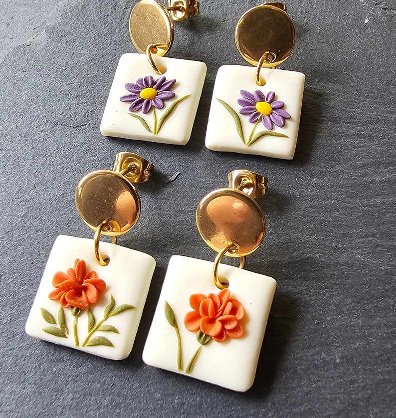 May include: Two pairs of drop earrings featuring gold-tone circular studs attached to white square pendants. One pair displays purple flowers with yellow centers, while the other pair features orange flowers with green stems and leaves on each square.