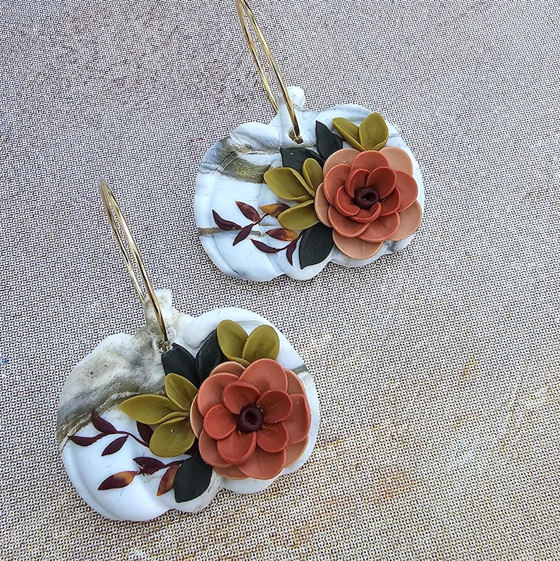 May include: A pair of white pumpkin-shaped earrings with gold hoops. Each earring features a marbled design, adorned with a layered orange flower, green and black leaves, and small brown leaf accents. The earrings are designed for pierced ears.