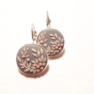 Peut inclure: Une paire de boucles d'oreilles pendantes rondes avec une base circulaire gris mat orn&eacute;e d'un motif en relief de feuilles beige clair et de petites baies roses. Elles sont mont&eacute;es sur un cadre en m&eacute;tal argent&eacute; avec des fermoirs dormeuses.