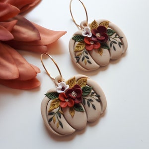 Pode incluir: Um par de brincos em forma de ab&oacute;bora bege com argolas douradas. Cada brinco &eacute; adornado com detalhes florais bord&ocirc;, brancos e amarelos e folhas verdes. Os brincos est&atilde;o sobre um fundo branco, com uma grande flor cor de ferrugem no canto superior esquerdo.