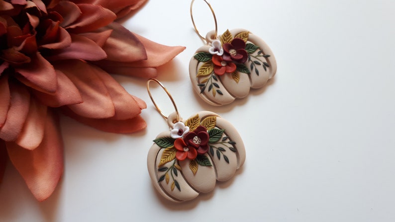 Pode incluir: Um par de brincos em forma de ab&oacute;bora bege com argolas douradas. Cada brinco &eacute; adornado com detalhes florais bord&ocirc;, brancos e amarelos e folhas verdes. Os brincos est&atilde;o sobre um fundo branco, com uma grande flor cor de ferrugem no canto superior esquerdo.