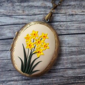 May include: Oval locket pendant with a gold-colored frame and a cream-colored center. The center features a cluster of yellow daffodils with orange centers and green stems. The locket hangs from a matching chain.