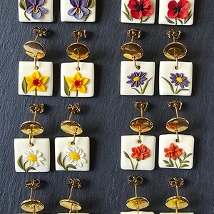May include: Twelve pairs of square white earrings featuring raised floral designs in various colors, including snowdrops, violets, poppies, daffodils, daisies, and roses. Each earring has a gold-toned circular stud top and a dangling square base.