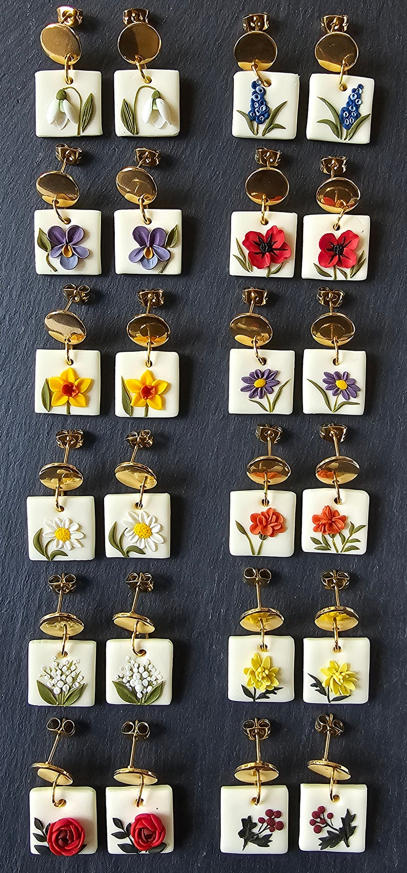 May include: Twelve pairs of square white earrings featuring raised floral designs in various colors, including snowdrops, violets, poppies, daffodils, daisies, and roses. Each earring has a gold-toned circular stud top and a dangling square base.