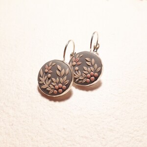 Peut inclure: Une paire de boucles d'oreilles rondes avec une base gris fonc&eacute; orn&eacute;e d'un motif floral et v&eacute;g&eacute;tal en relief beige clair et de petites baies roses. Les disques circulaires sont mont&eacute;s sur une monture en m&eacute;tal argent&eacute; avec des fermoirs dormeuses.