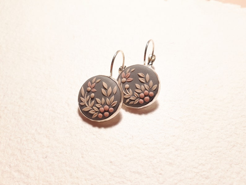 Peut inclure: Une paire de boucles d'oreilles rondes avec une base gris fonc&eacute; orn&eacute;e d'un motif floral et v&eacute;g&eacute;tal en relief beige clair et de petites baies roses. Les disques circulaires sont mont&eacute;s sur une monture en m&eacute;tal argent&eacute; avec des fermoirs dormeuses.