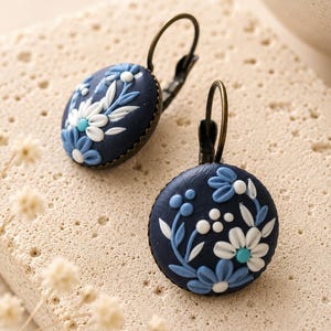 May include: A pair of round drop earrings featuring a dark navy blue base with raised floral motifs in white, light blue, and turquoise. The circular discs are set in an antique bronze-toned metal frame with matching lever-back ear wires.