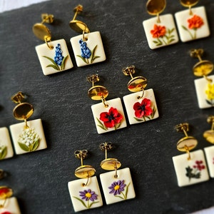 May include: A collection of square-shaped earrings featuring various floral designs on a white base. Each pair includes gold-toned metal studs with dangling square pendants decorated with sculpted flowers in blue, orange, red, white, and purple.