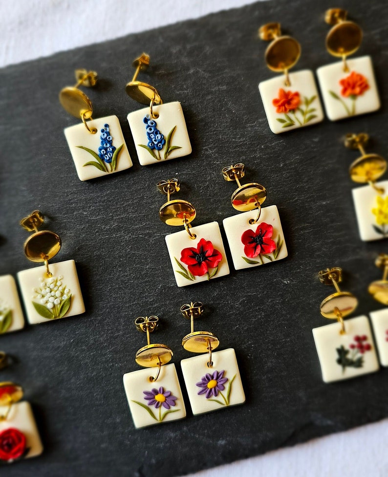 May include: A collection of square-shaped earrings featuring various floral designs on a white base. Each pair includes gold-toned metal studs with dangling square pendants decorated with sculpted flowers in blue, orange, red, white, and purple.