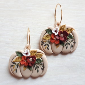 Pode incluir: Um par de brincos em forma de ab&oacute;bora bege com argolas douradas. Cada brinco &eacute; adornado com um grupo de flores vermelhas, laranja e brancas, juntamente com folhas verdes e amarelas. Os brincos s&atilde;o projetados com um tema de colheita de outono.