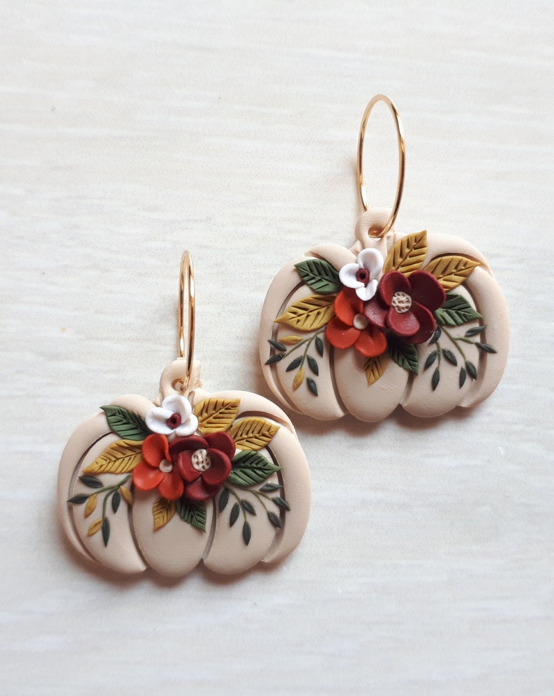 Pode incluir: Um par de brincos em forma de ab&oacute;bora bege com argolas douradas. Cada brinco &eacute; adornado com um grupo de flores vermelhas, laranja e brancas, juntamente com folhas verdes e amarelas. Os brincos s&atilde;o projetados com um tema de colheita de outono.