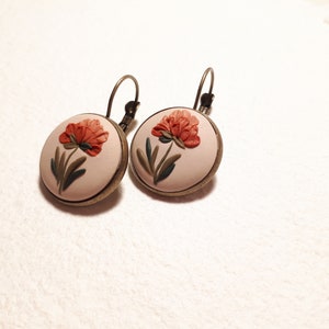 May include: A pair of round earrings featuring a cream-colored base with a raised, sculpted orange flower and green leaves. The circular design is set in a dark bronze-toned metal frame and includes a matching bronze-toned leverback closure.