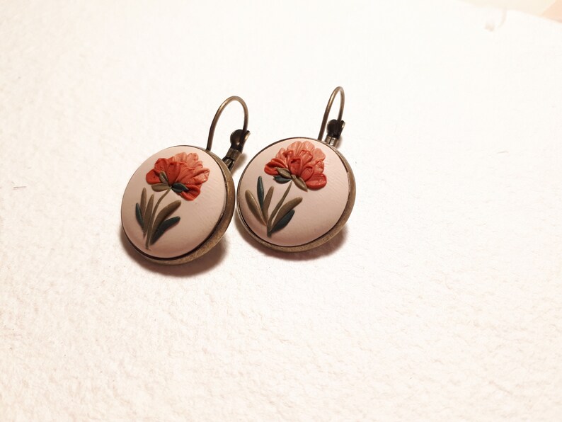 May include: A pair of round earrings featuring a cream-colored base with a raised, sculpted orange flower and green leaves. The circular design is set in a dark bronze-toned metal frame and includes a matching bronze-toned leverback closure.