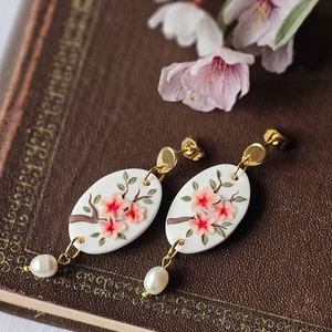 May include: A pair of white ceramic earrings with gold accents. Each earring features a delicate branch with pink blossoms. The earrings are hanging from a book with a brown leather cover.