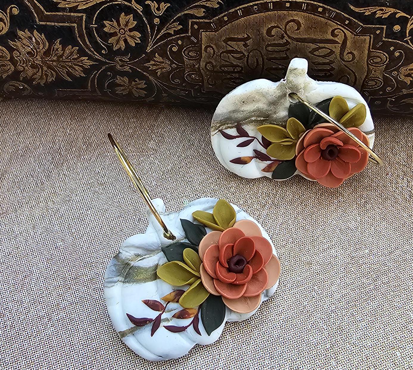 May include: Two decorative pumpkin-shaped earrings. The earrings are white with gold marbling and adorned with orange and green floral accents. Each earring has a gold-colored hoop.