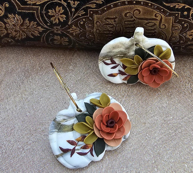 May include: Two decorative pumpkin-shaped earrings. The earrings are white with gold marbling and adorned with orange and green floral accents. Each earring has a gold-colored hoop.