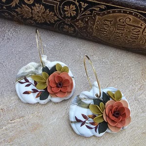 May include: A pair of white pumpkin-shaped earrings with gold hoops. Each earring features a 3D floral design with orange and green leaves. The earrings have a marbled effect and are set against a neutral background.