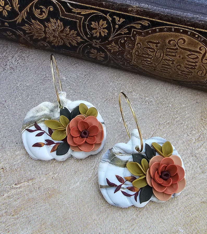 May include: A pair of white pumpkin-shaped earrings with gold hoops. Each earring features a 3D floral design with orange and green leaves. The earrings have a marbled effect and are set against a neutral background.