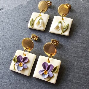 May include: Two pairs of drop earrings featuring gold-tone circular studs attached to square off-white pendants. One pair displays white snowdrop flowers with green stems, while the other pair features purple pansies with yellow centers and green leaves.