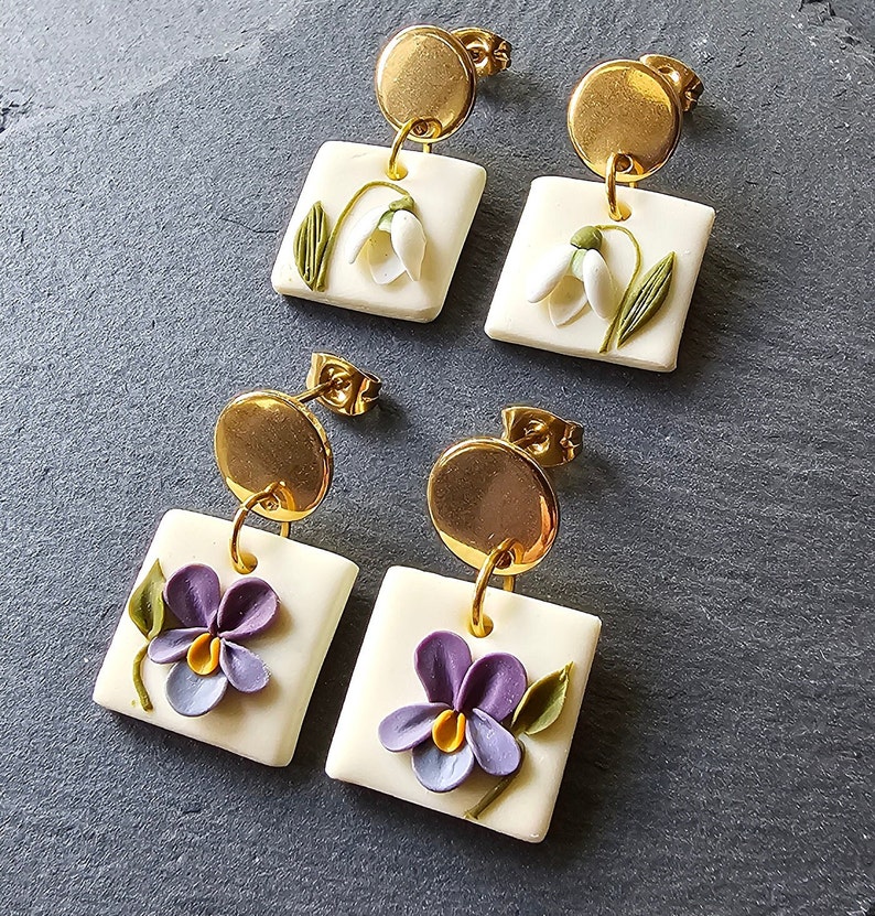 May include: Two pairs of drop earrings featuring gold-tone circular studs attached to square off-white pendants. One pair displays white snowdrop flowers with green stems, while the other pair features purple pansies with yellow centers and green leaves.