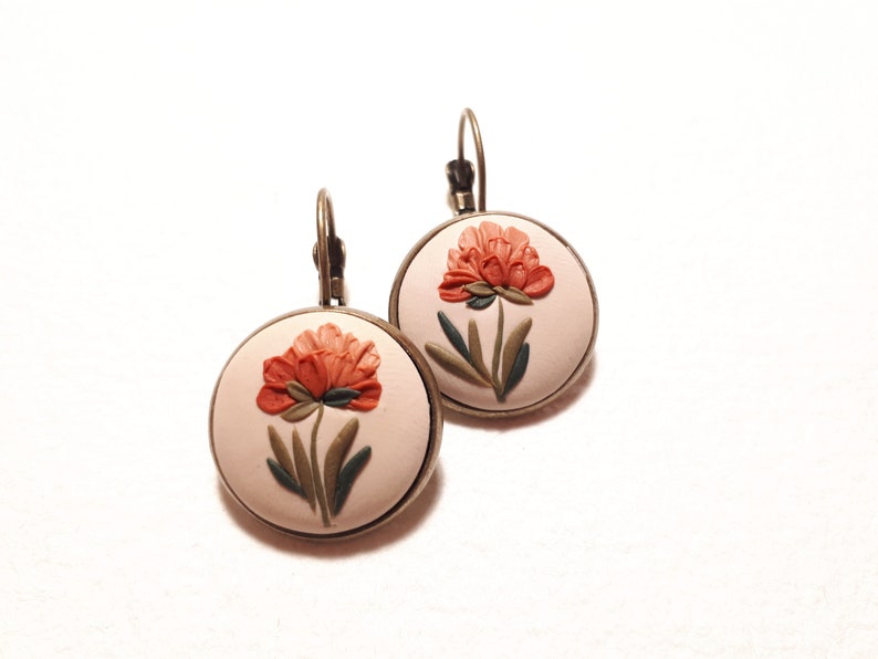 May include: A pair of round drop earrings featuring a light beige background with a raised, sculpted orange flower and dark green leaves. The circular design is set in a bronze-toned metal frame with a matching lever-back closure mechanism.