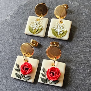 May include: Two pairs of drop earrings featuring gold-tone circular studs connected to square off-white pendants. One pair displays embossed white lily of the valley flowers with green leaves, while the other pair features a raised red rose with green leaves.