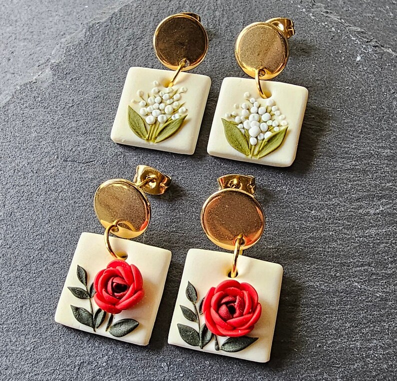 May include: Two pairs of drop earrings featuring gold-tone circular studs connected to square off-white pendants. One pair displays embossed white lily of the valley flowers with green leaves, while the other pair features a raised red rose with green leaves.