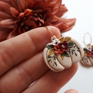 Pode incluir: Um par de brincos bege em forma de ab&oacute;bora com argolas douradas. Cada brinco &eacute; adornado com pequenas flores coloridas e folhas verdes. Um acess&oacute;rio com tema de outono.