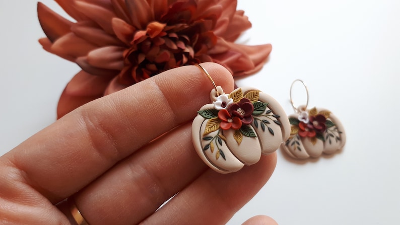 Pode incluir: Um par de brincos bege em forma de ab&oacute;bora com argolas douradas. Cada brinco &eacute; adornado com pequenas flores coloridas e folhas verdes. Um acess&oacute;rio com tema de outono.