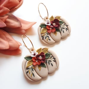 Pode incluir: Um par de brincos em forma de ab&oacute;bora bege com argolas douradas. Cada brinco &eacute; adornado com um aglomerado de flores vermelhas, brancas e amarelas e folhas verdes. Os brincos est&atilde;o sobre um fundo branco.