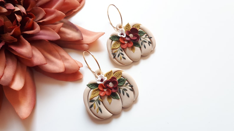 Pode incluir: Um par de brincos em forma de ab&oacute;bora bege com argolas douradas. Cada brinco &eacute; adornado com um aglomerado de flores vermelhas, brancas e amarelas e folhas verdes. Os brincos est&atilde;o sobre um fundo branco.