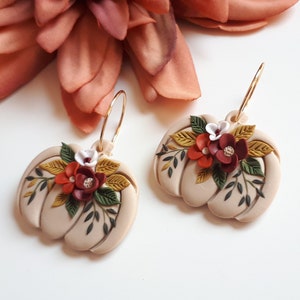Pode incluir: Um par de brincos bege em forma de ab&oacute;bora com argolas douradas. Cada brinco &eacute; adornado com um aglomerado de flores vermelhas, brancas e bord&ocirc;, juntamente com folhas verdes e amarelas, criando um design outonal.