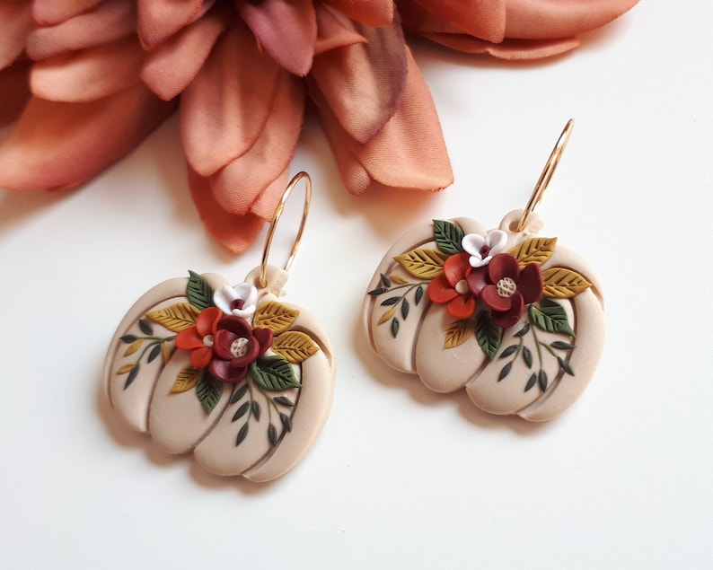 Pode incluir: Um par de brincos bege em forma de ab&oacute;bora com argolas douradas. Cada brinco &eacute; adornado com um aglomerado de flores vermelhas, brancas e bord&ocirc;, juntamente com folhas verdes e amarelas, criando um design outonal.