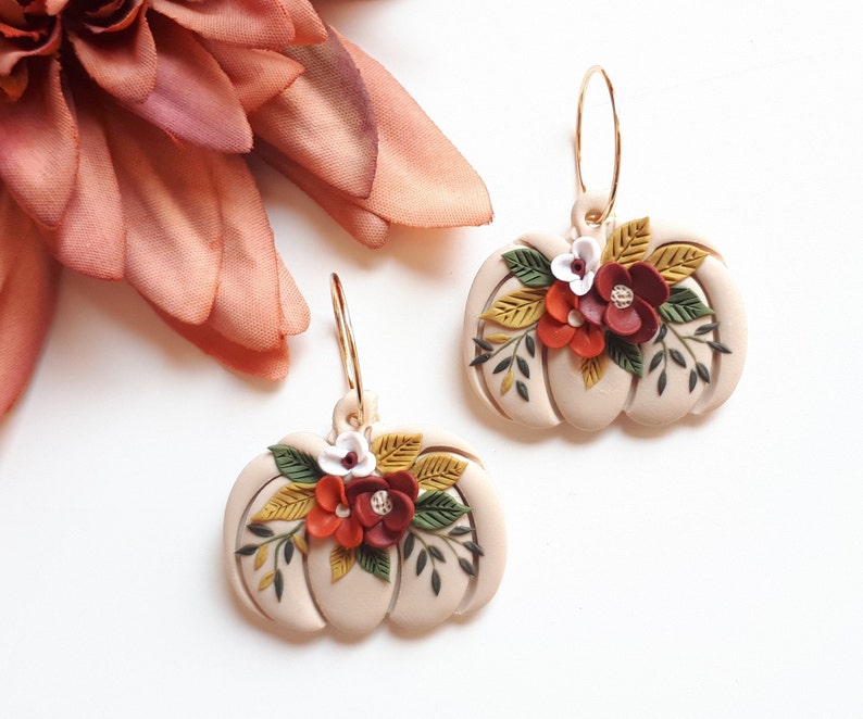 Pode incluir: Um par de brincos em forma de ab&oacute;bora bege com argolas douradas. Cada brinco &eacute; adornado com um aglomerado de flores vermelhas, brancas e amarelas e folhas verdes. Os brincos est&atilde;o em um fundo branco.